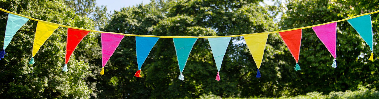 3m string of colourful cotton bunting with green trees and sunshine background. Line is yellow and flags are light blue, yellow, red, pink, blue, turquoise with contrast tassles.