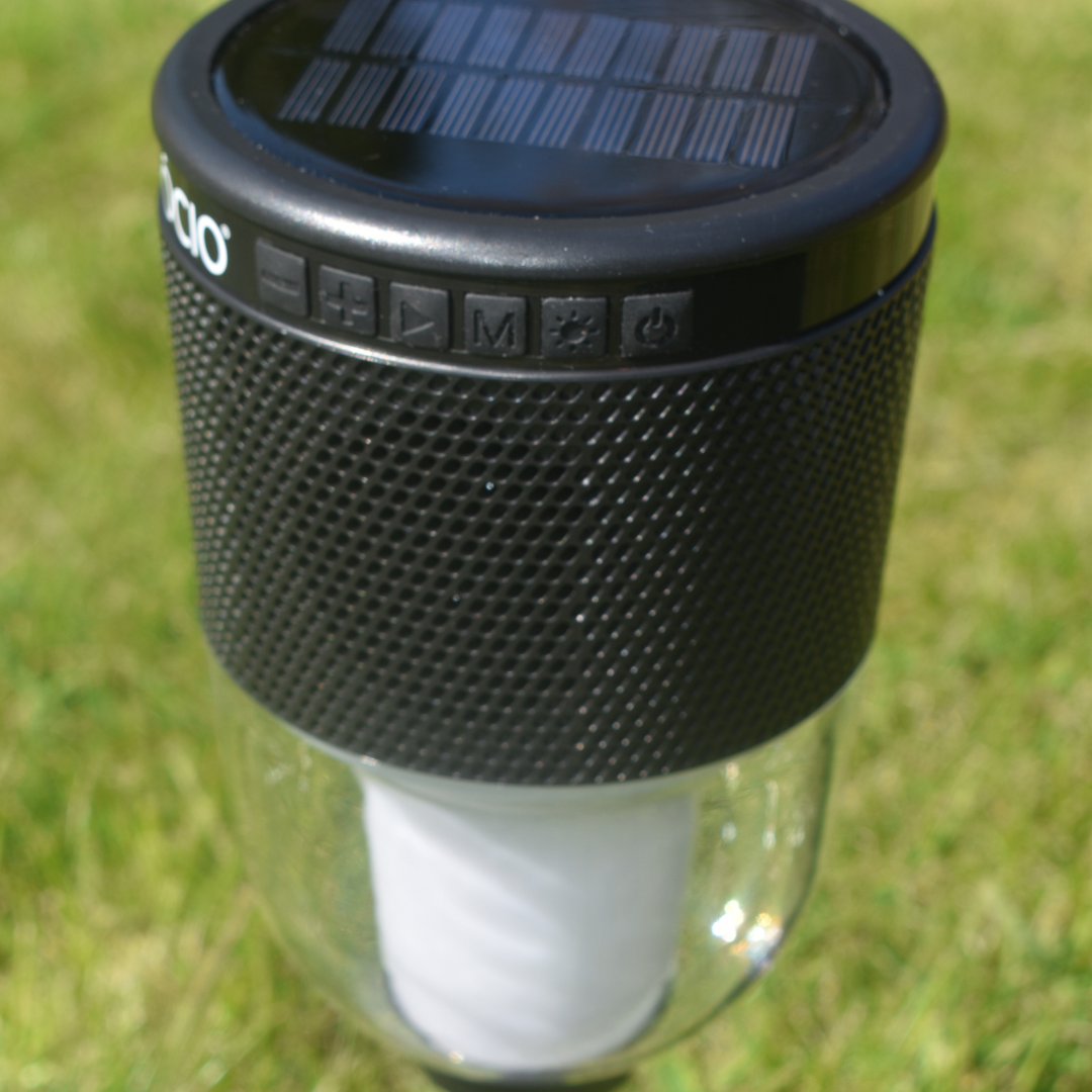 close up colour change lantern and speaker with ground spike pictured green grass background, black solar panel on top and speaker control.
