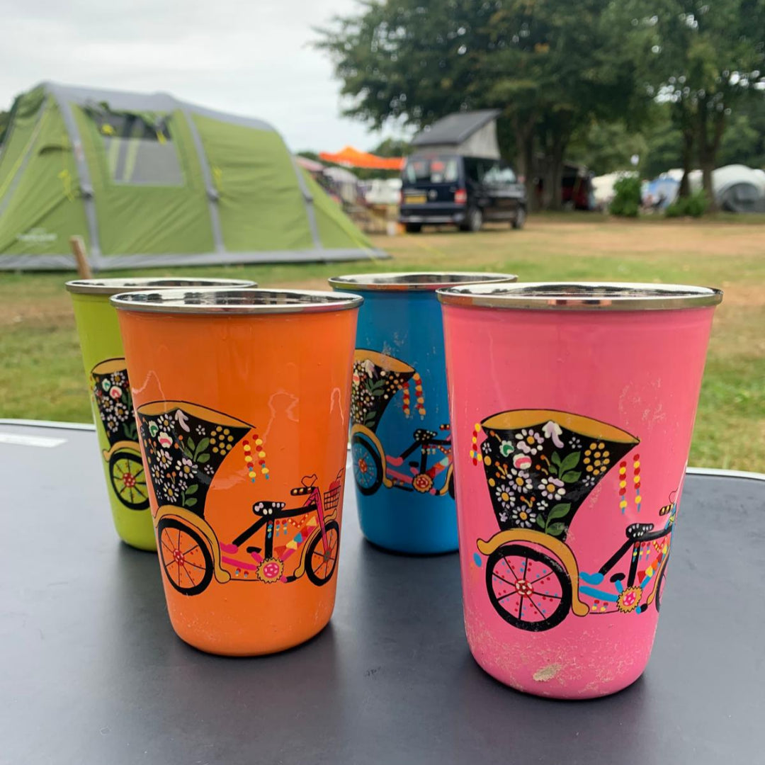 stainless steel enamelled wine tumblers with hand painted colourful rickshaw designs on a camping table with tents and trees in the background.