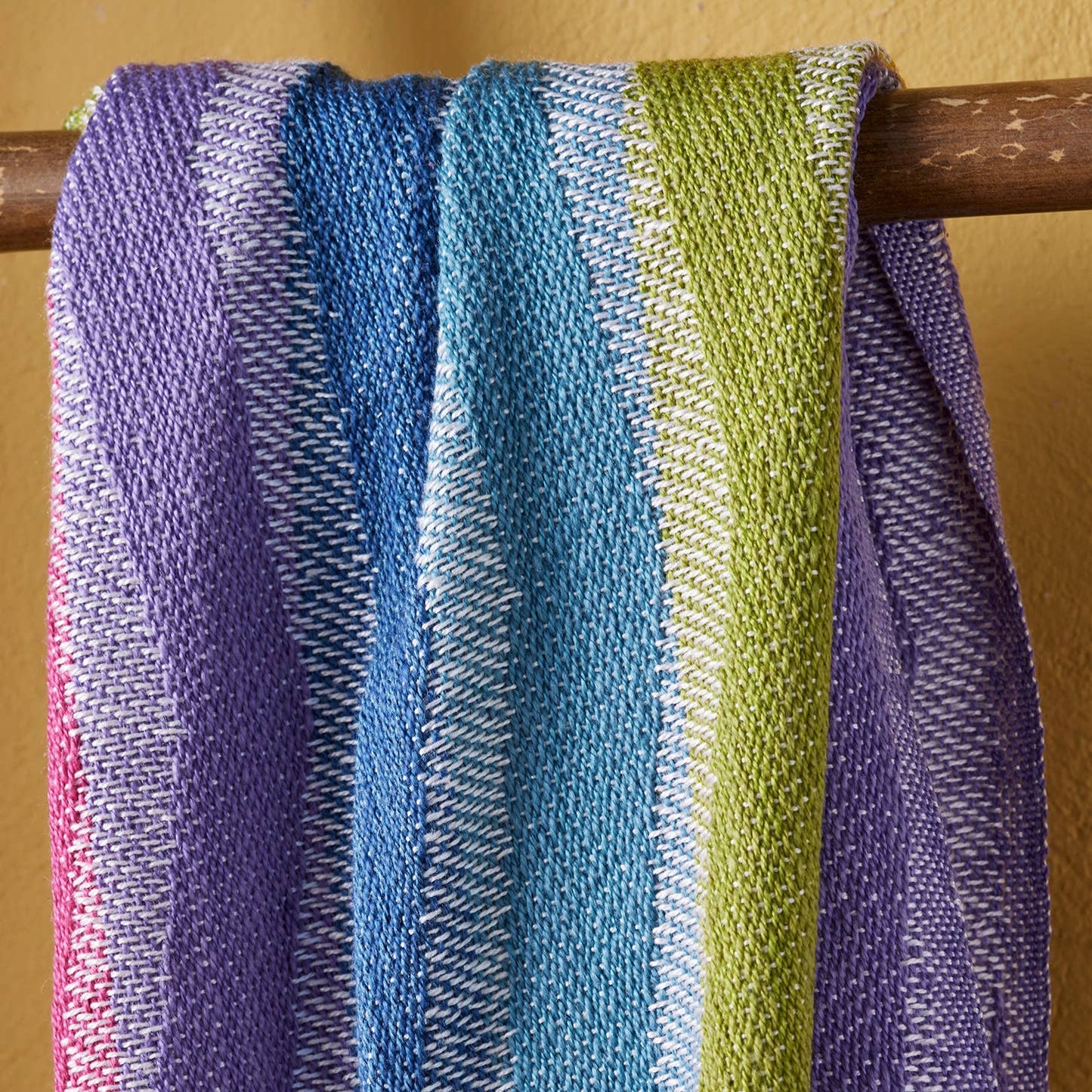 Multicolored striped throw on a wooden rod against a yellow wall.
