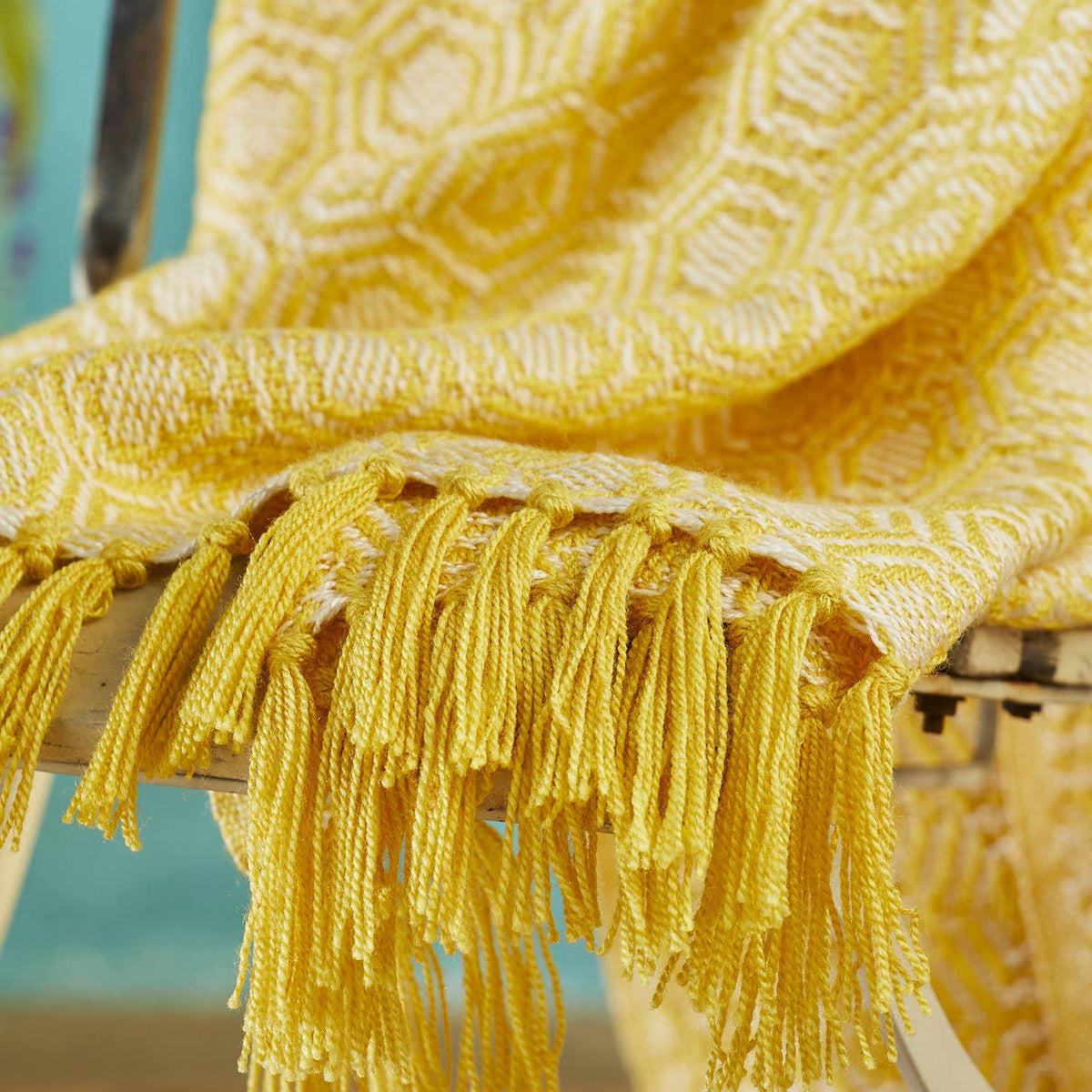 A close-up of a yellow woven throw blanket draped over a chair, featuring a geometric pattern and long fringe tassels.