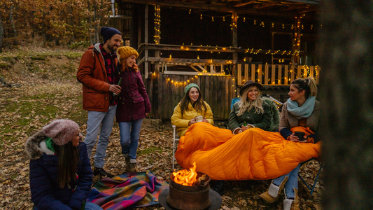 a group of friends sitting around a camp firte with warm coats, hats and blankets with a wooden hut and fairy lights in the background