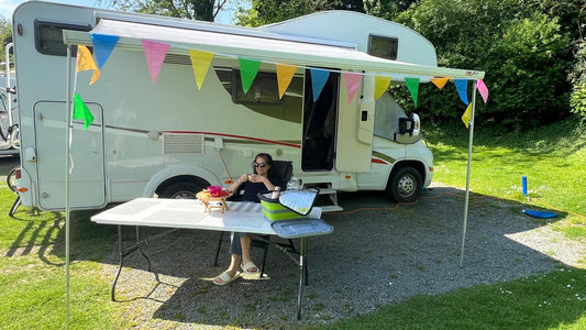 white motorhome with awning and bunting. Lady sipping coffee under the awning at large white table.