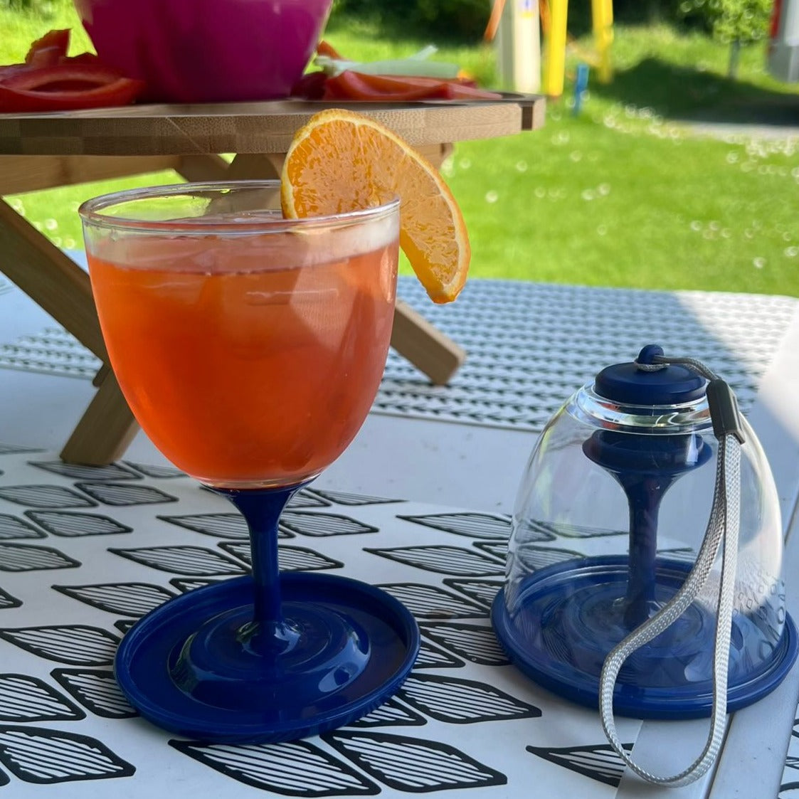 Two clear plastic wine glasses with royal blue stem and wide base. Left glass filled with aperol spritz with orange slice. Right glass packed away with carry lanyard.