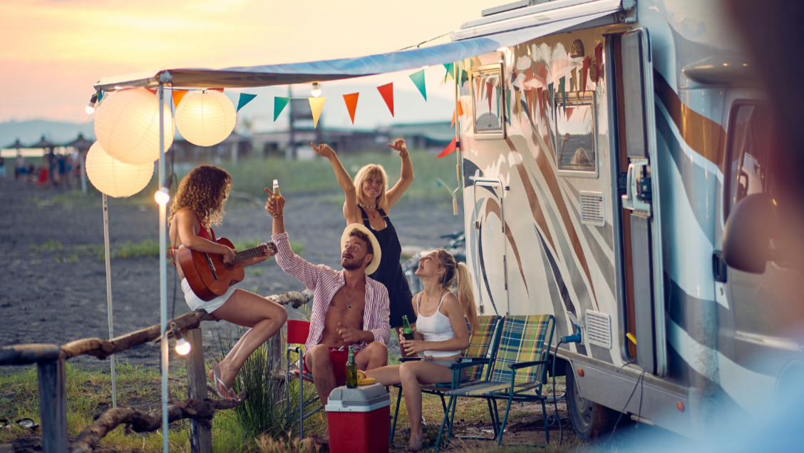 three females and a male pitched up next to a beach, sitting outside a motorhome, sharing drinks, laughing, dancing and singing. Awning is decorated with bunting and lighting.