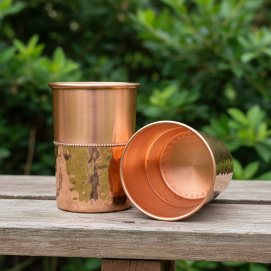 Two copper tumblers on a wooden surface with a green foliage background