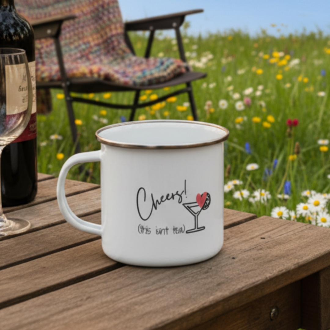 White mug with 'Cheers! (this isn't tea)' text on a wooden table with a bottle and glass in a field.