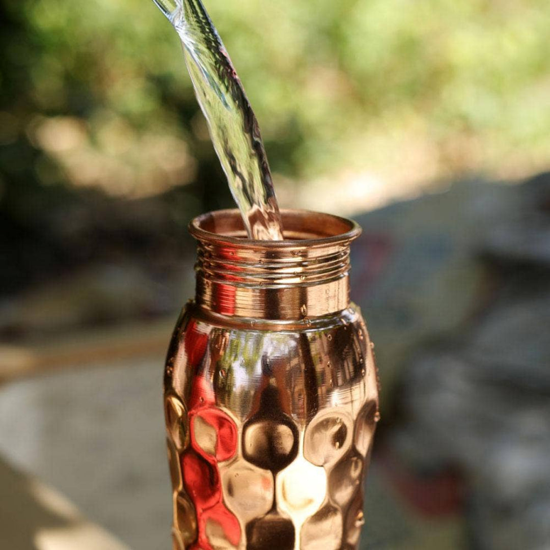 Copper hammered water bottle with water pouring into it. Outdoors blurred background.