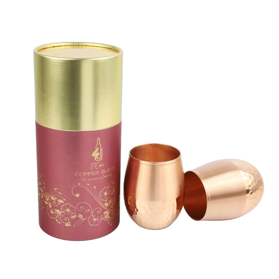 A copper tumbler glass shown with its gold-and-deep-red cylindrical packaging tube on a light background. One cup is upright and one is tipped on its side, both showing a hammered metallic copper finish.
