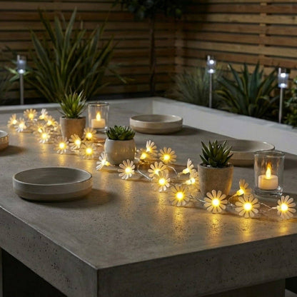 daisy shaped string lights lying across an outdoor table with plants and a fence in the background.