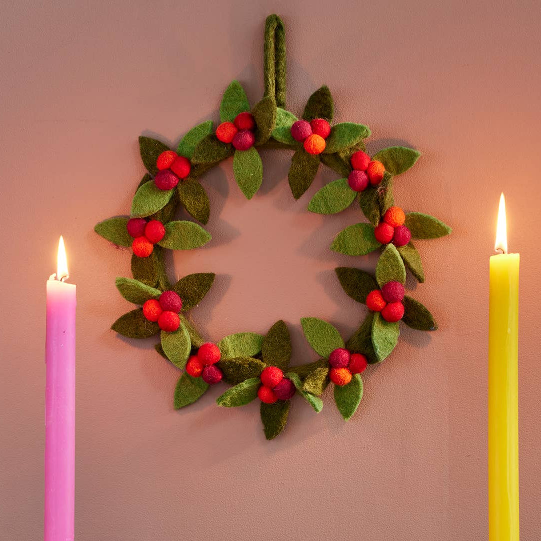 Two colorful candlesticks with pink and yellow candles in front of a green and red berry decorative wreath