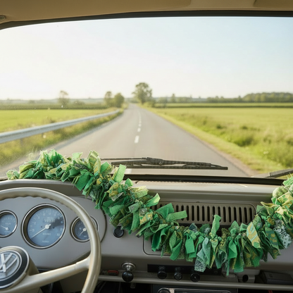 A green fabric garland made of recycled saris is draped across the dashboard of a vanr. The van is driving on a country road with green fields on both sides.