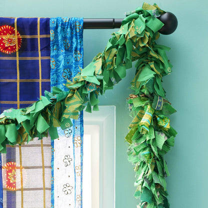 A green fabric garland made of mixed green sari fabrics, draped over a black curtain rod, next to patterned blue and white curtains against a light teal wall.