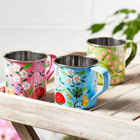 Three colorful hand-painted stainless steel camping mugs with floral designs on a wooden surface.