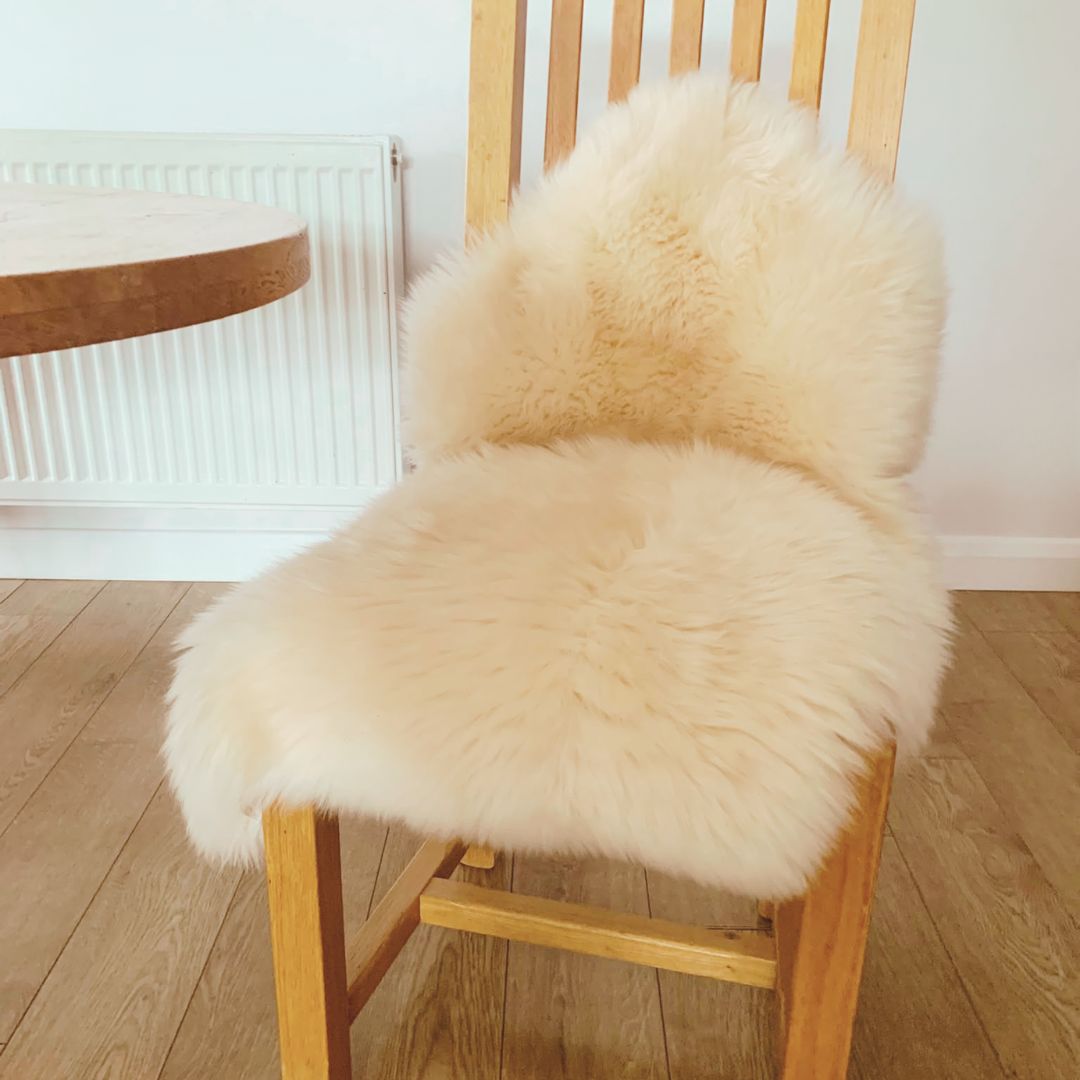 A natural coloured sheepskin draped over a light wood dining chair in a bright room with a wooden floor and white wall.