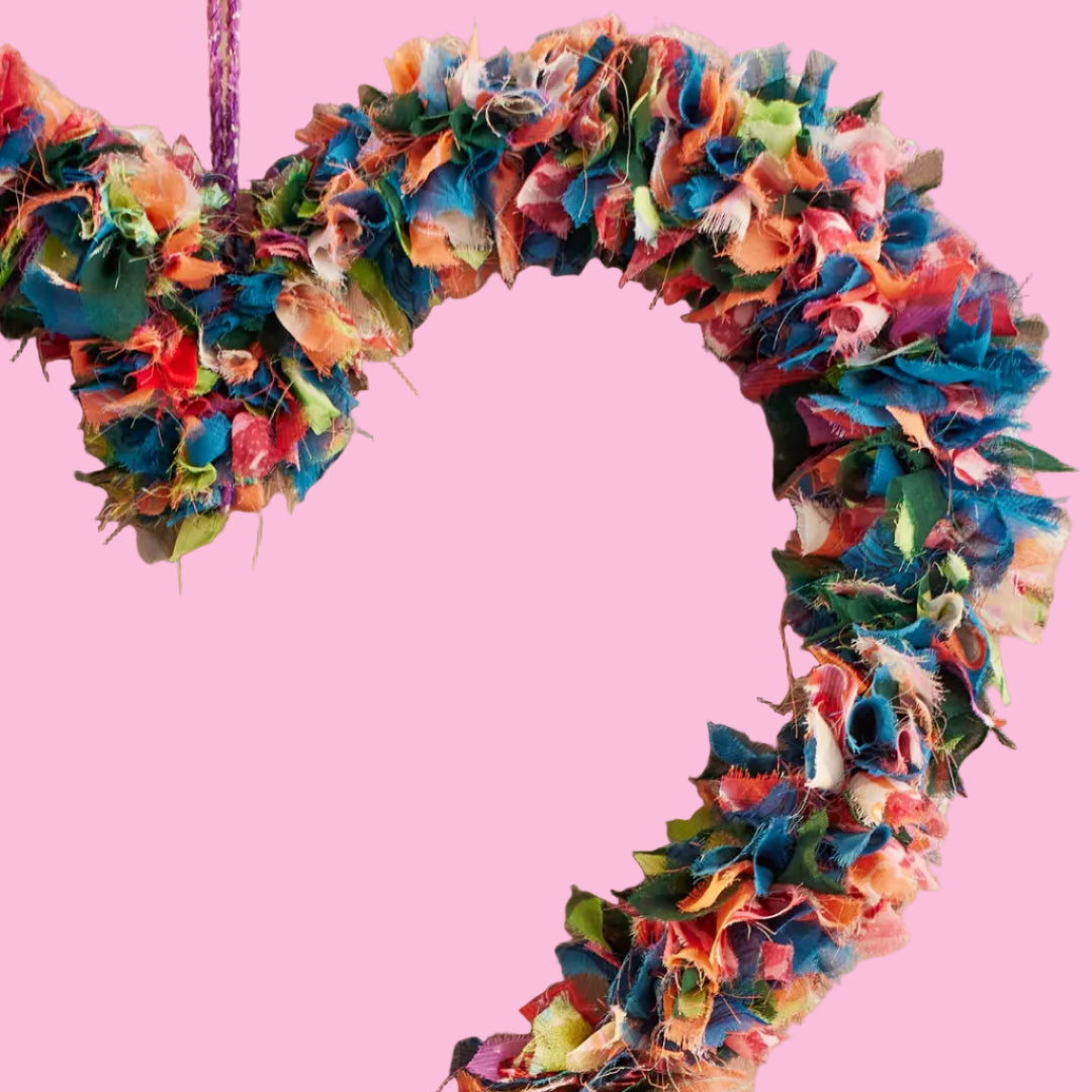 A close-up of a heart-shaped fabric wreath made from multicolored sari remnants, hanging against a pink background. Only a quarter of the wreath is visible.
