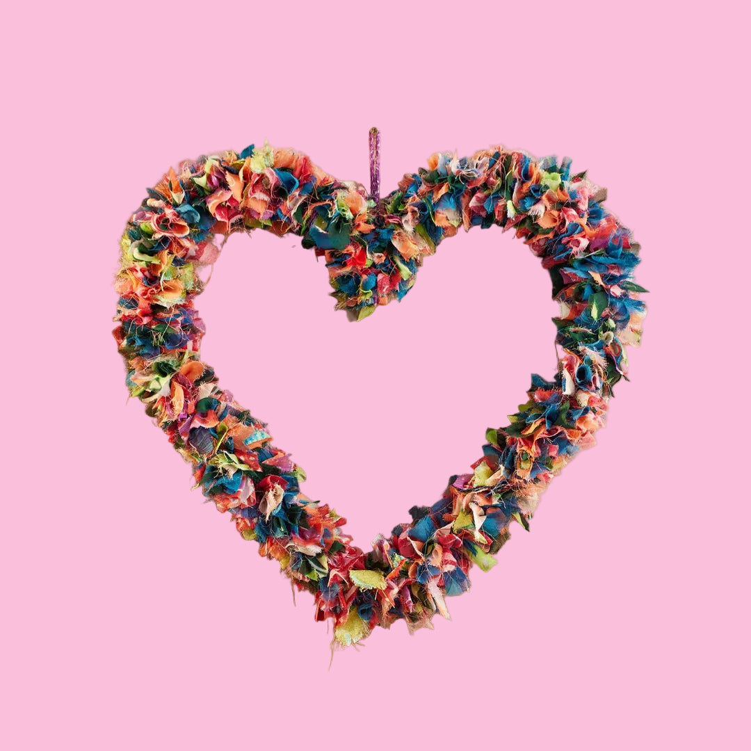 A heart-shaped fabric wreath made from colorful sari remnants on a pink background