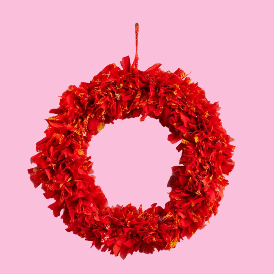 A large red wreath made of recycled sari fabrics hanging on a red string on a pink background