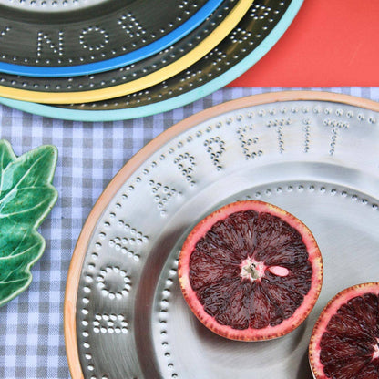 A styled tabletop scene showing a steel plate with orange trim and embossed lettering and dot patterns, holding two cut halves of red blood orange. Other decorative plates are stacked in the background on a blue-and-white gingham cloth, with a green ceramic leaf and a red surface visible.