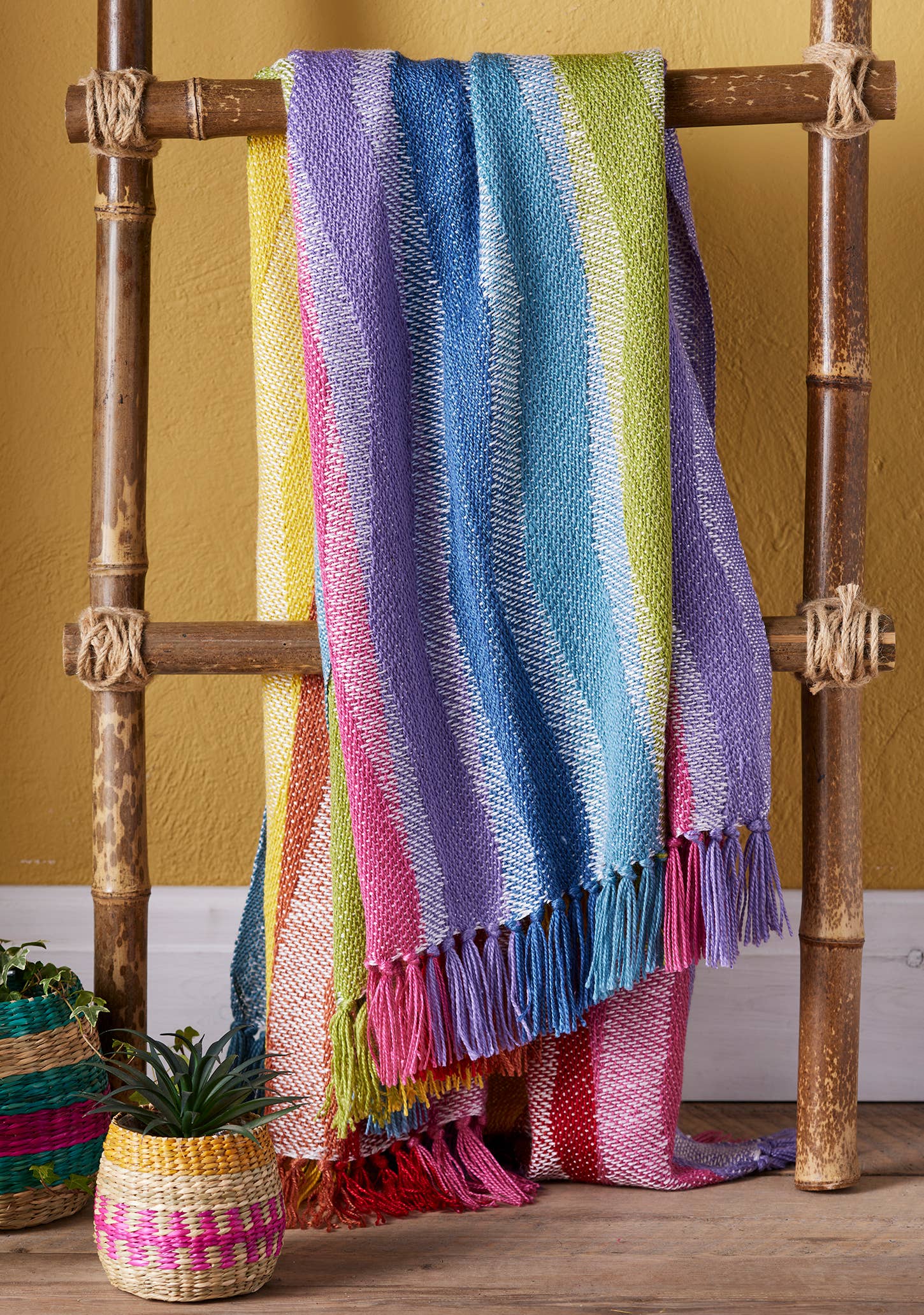 Colourful striped throw made from recycled bottles draped over a wooden rack against a yellow wall.