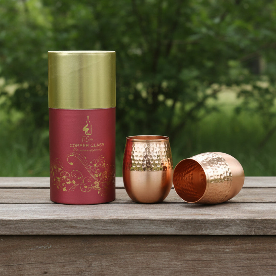 A copper tumbler glass shown with its gold-and-deep-red cylindrical packaging tube on wooden table with trees beyond. One cup is upright and one is tipped on its side, both showing a hammered metallic copper finish.