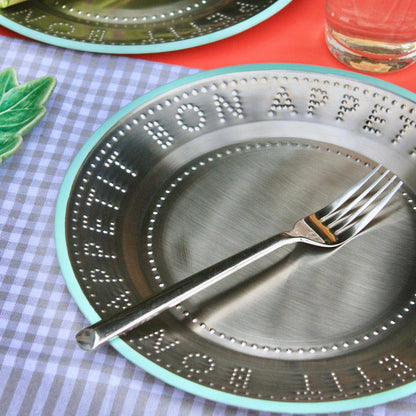 A close-up of a metal plate with a mint green trim, embossed text around the edge, and a silver fork resting across the center, placed on a checkered tablecloth.