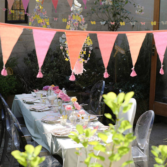 Decorated outdoor table with pink and peach bunting, floral centerpieces, and greenery.