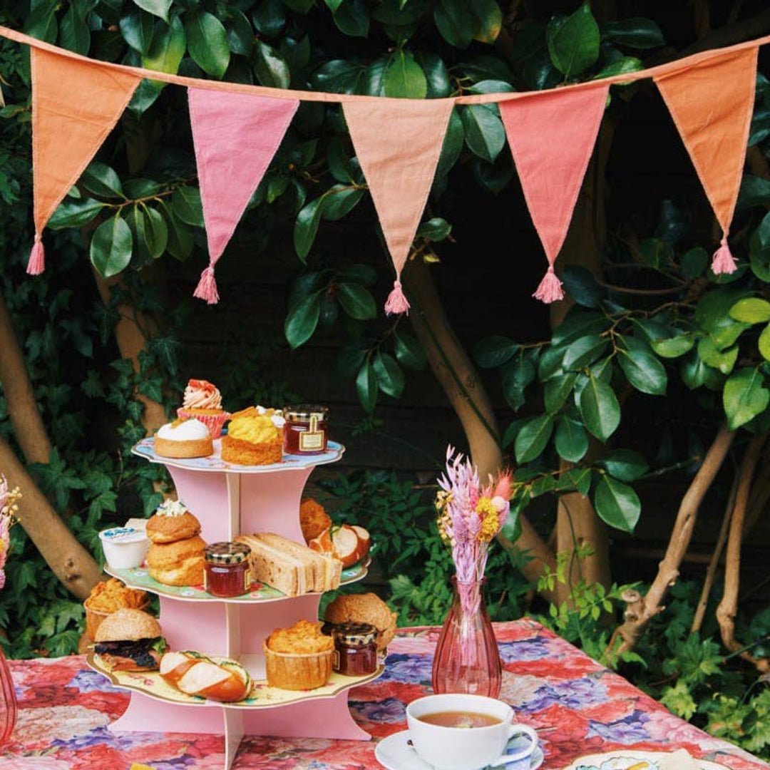 Outdoor tea party setup with a floral tablecloth, pink pastel bunting, and a three-tiered cake stand.
