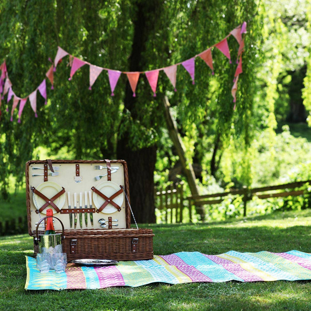 Open picnic basket with cutlery on a colorful blanket under a tree with pink tassel bunting