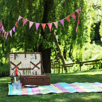Open picnic basket with cutlery on a colorful blanket under a tree with pink tassel bunting