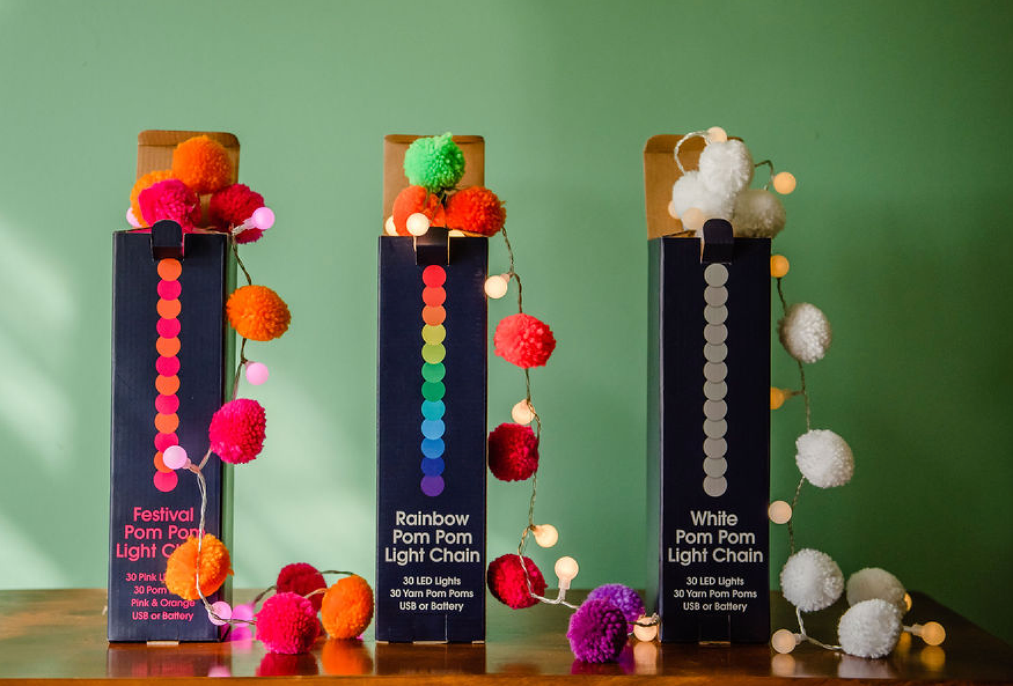 Three boxes of pom-pom light chains with colorful pom-poms on a wooden surface against a green background.