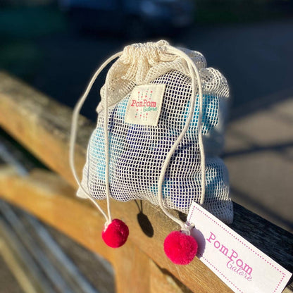 A small mesh drawstring pouch with white cords and red pom-poms, holding a rainbow pompom string, resting on a wooden fence outdoors. A tag reads 'PomPom Galore' and a small label is visible on the pouch.