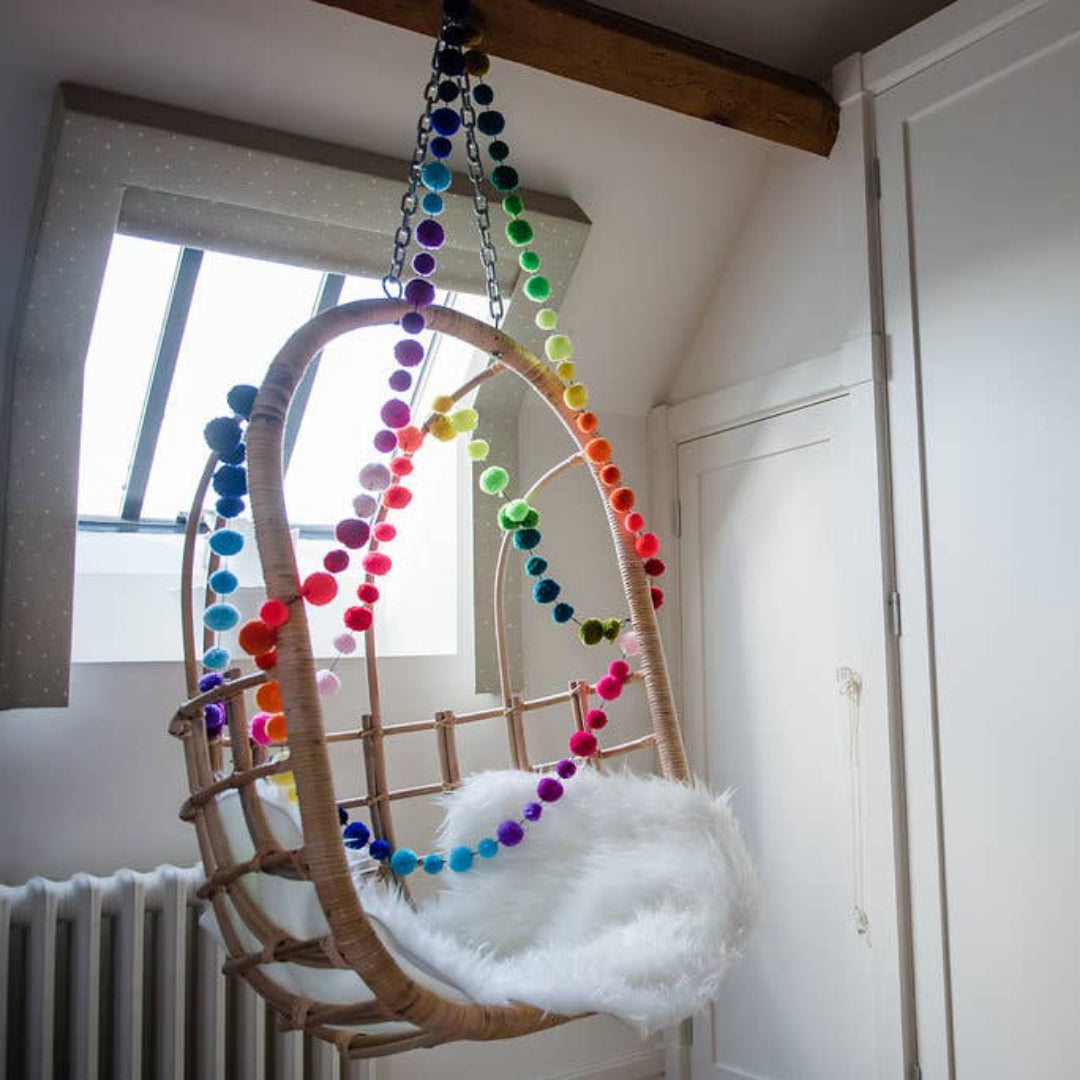 A hanging rattan baby swing chair with a white faux fur cushion, decorated with colorful rainbow pom-pom garlands, set in a bright room with white walls and a window.