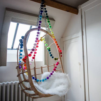A hanging rattan baby swing chair with a white faux fur cushion, decorated with colorful rainbow pom-pom garlands, set in a bright room with white walls and a window.