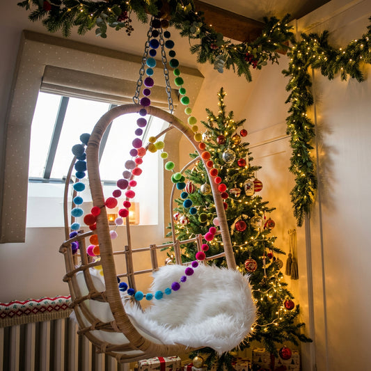A hanging rattan baby swing chair with a white faux fur cushion, decorated with colorful rainbow pom-pom garlands, set in a bright room with white walls and a window.