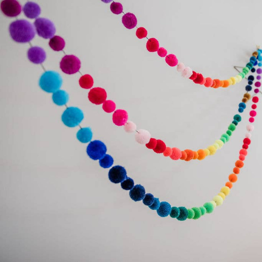 A colourful pom-pom string with three curved rows of rainbow-colored fuzzy balls, hanging against a plain white wall next to a silver star ornament.