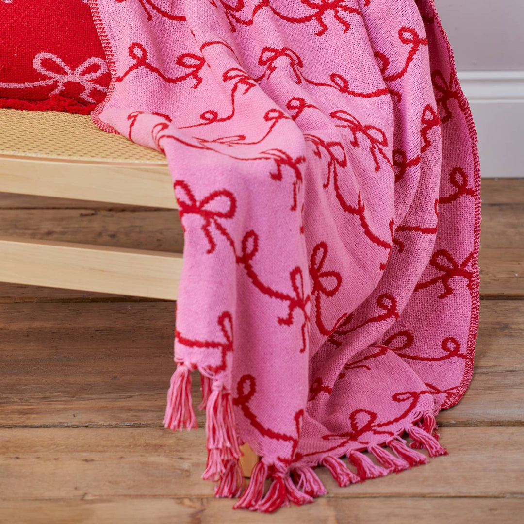 A pink woven throw blanket with red embroidered bow patterns and fringe, draped over a light wood bench next to a red matching pillow on a wooden floor.