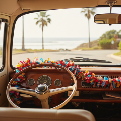 A vintage-style campervan interior with a wood dashboard and steering wheel, decorated with a colorful fabric garlands draped across it, with a scenic coastal road and palm trees visible through the windshield.