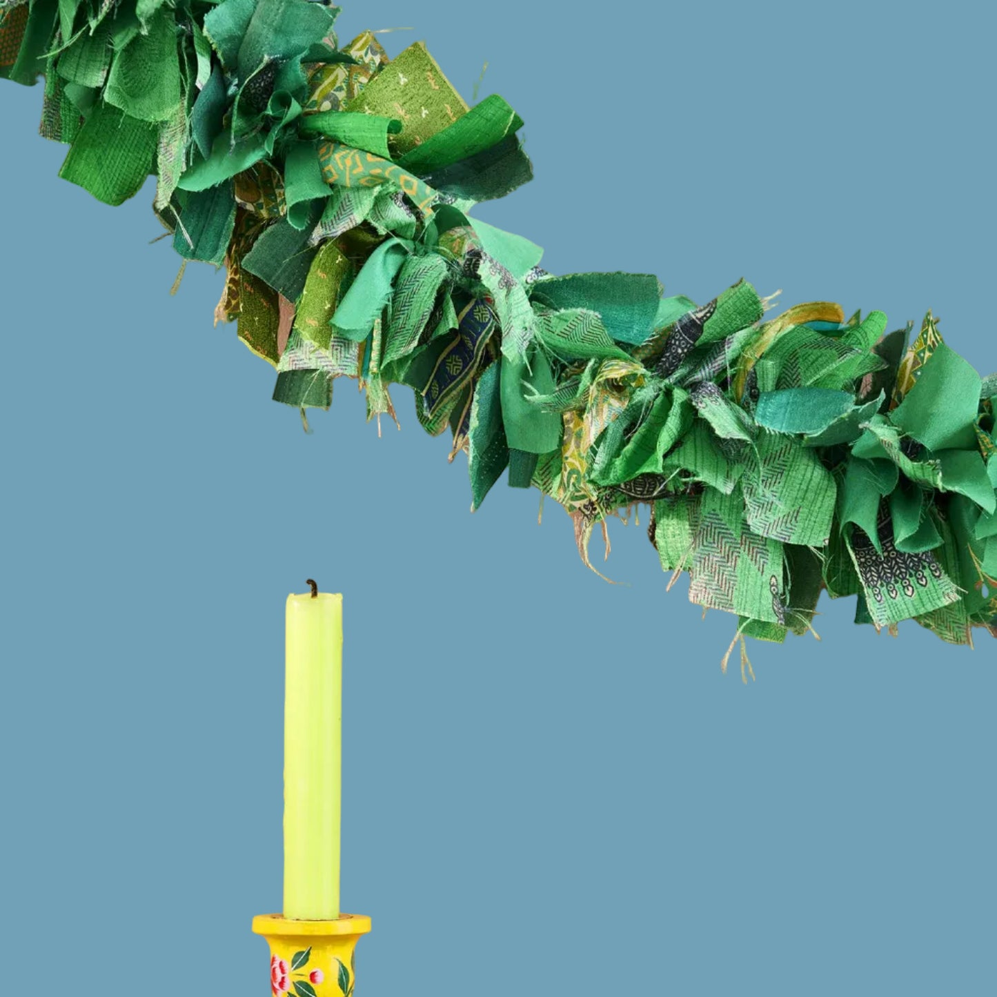 A festive green fabric garland hangs on a blue wall above a yellow candle and candlestick holder