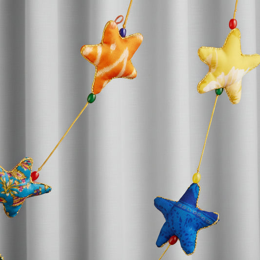 Colorful fabric star garland hanging against a warm brown wooden door. The stars are in orange, yellow, blue, pink, and a multicolor floral pattern, strung on thin yellow cords with small beads.