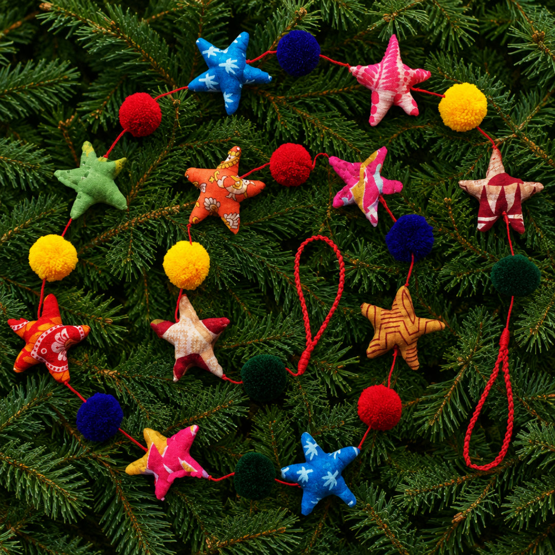 A colorful fabric garland featuring stars and pompoms on a coloured string a green pine branch background.