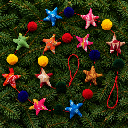 A colorful fabric garland featuring stars and pompoms on a coloured string a green pine branch background.