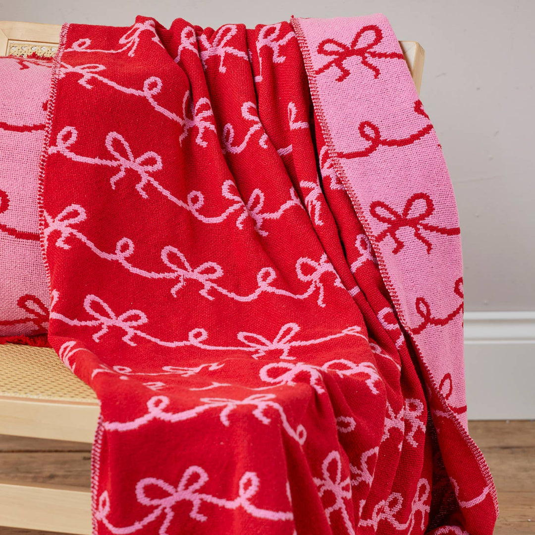 A red and pink woven throw blanket with a repeating bow pattern, draped over a light wood chair on a wooden floor.