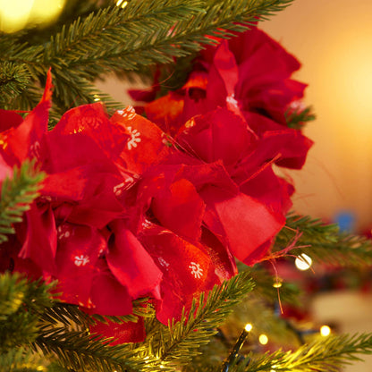 A close-up of a Christmas tree decorated with large red fabric tinsel, green pine branches, and warm yellow string lights in a softly blurred indoor setting.