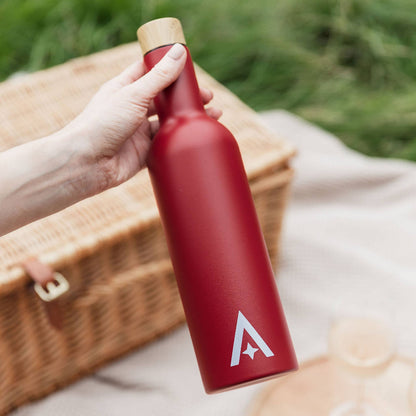 Red insulated wine bottle with a cork cap held by a hand on a picnic blanket with a basket
