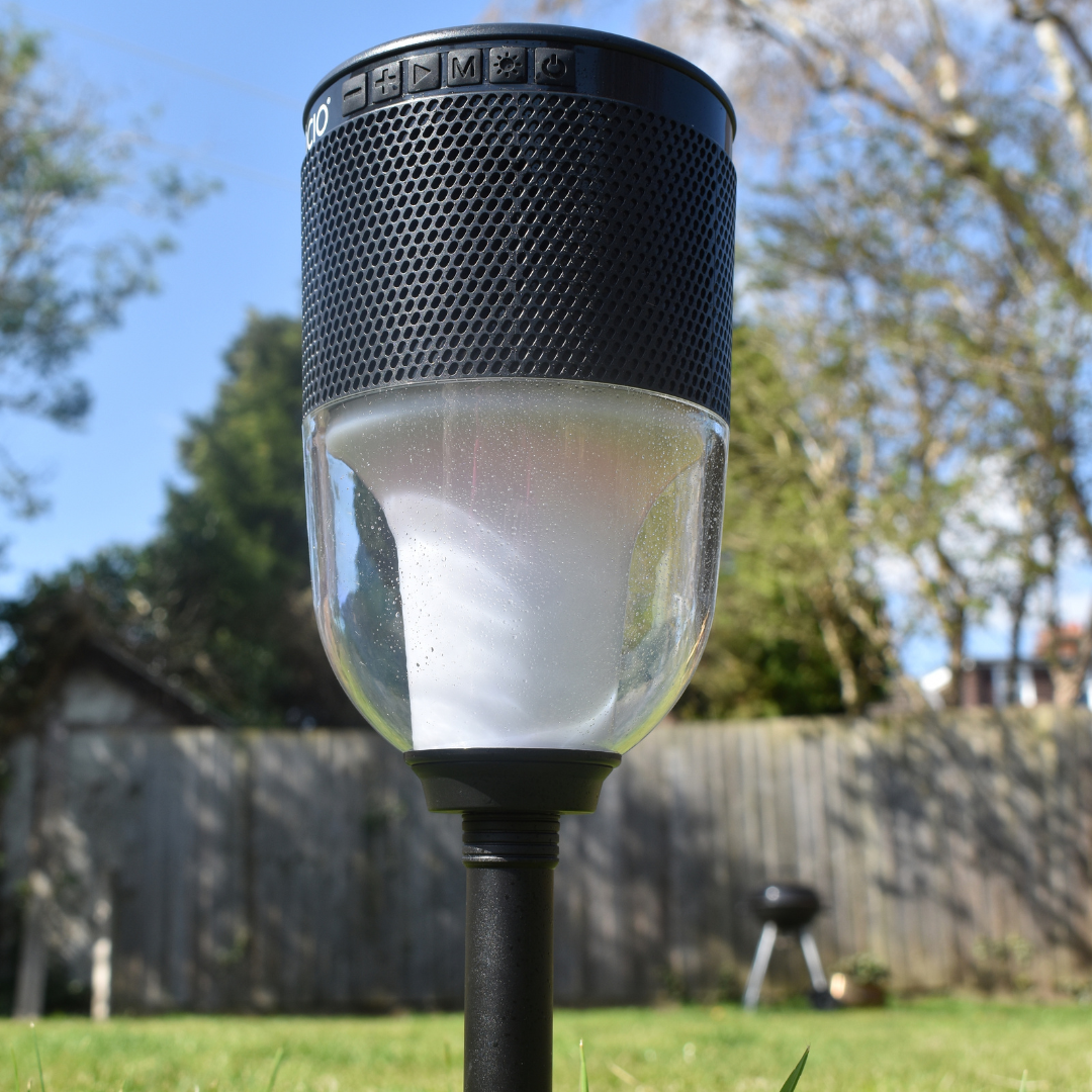 colour change lantern and speaker with ground spike pictured with garden background, grey fence, trees and black barbecue.