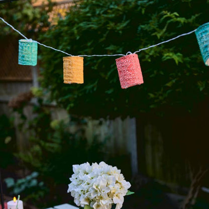 Outdoor table setting with flowers, candles, and colourful string lights against a greenery background.