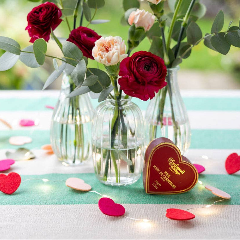 Three small glass vases with red, pink, and white flowers on a tablecloth with heart string light decorations