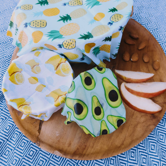 Small, medium and large bowls covered with wax food wraps on a wooden plate with sliced apples on a blue patterned background
