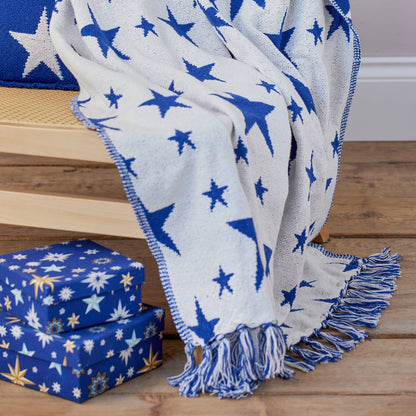 A soft woven throw blanket draped over a wooden chair, featuring a blue and white star pattern with fringed edges. Matching star-printed boxes sit on the floor below.